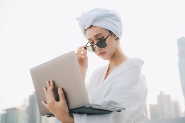 Güneş gözlüğü takan beyaz bornozlu genç bir kadın dizüstü bilgisayarla çalışıyor. Evde çalışıyorum. Duş aldıktan sonra çalışıyorum..