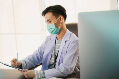 Maskeli yakışıklı Asyalı Taylandlı doktor çalışma masasında çalışıyor. Doktor, Covid-19 virüsünü tedavi edecek aşı üzerinde bilgisayar araştırması yapıyor..
