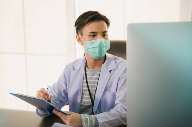 Maskeli yakışıklı Asyalı Taylandlı doktor çalışma masasında çalışıyor. Doktor, Covid-19 virüsünü tedavi edecek aşı üzerinde bilgisayar araştırması yapıyor..