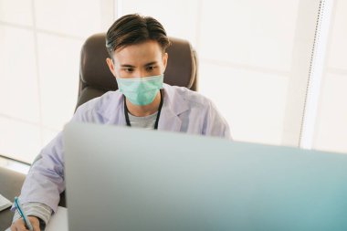 Maskeli yakışıklı Asyalı Taylandlı doktor çalışma masasında çalışıyor. Doktor, Covid-19 virüsünü tedavi edecek aşı üzerinde bilgisayar araştırması yapıyor..