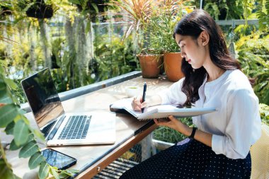 Çiçek ve ağaçlarla dolu bir bahçede deftere yazan kadın..