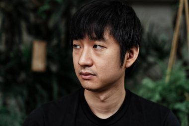 Siyah tişörtlü bir adamın, arka plan olarak sağ tarafına ve bahçesine bakarken yakın plan fotoğrafı..