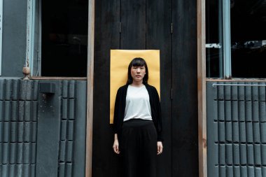 Siyah beyaz elbiseli bir kadın sarı kağıtlı siyah kapının önünde duruyor..