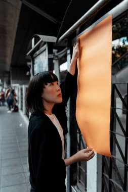 Siyah beyaz giysili bir kız kaldırım kenarındaki afişe turuncu kağıt yapıştırıyor..