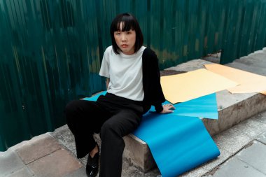 Siyah beyaz kumaşlı kadın yeşil duvarın yanında mavi, sarı ve turuncu kağıtlarla kayanın üzerinde oturuyor..