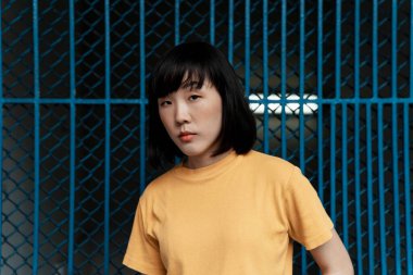 Turuncu tişörtlü bir kadın mavi çitin önünde heykel gibi duruyor..