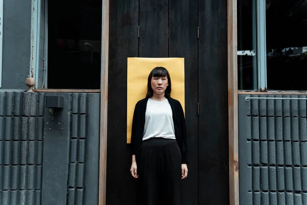 Siyah beyaz elbiseli bir kadın sarı kağıtlı siyah kapının önünde duruyor..