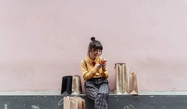 Милая Девушка Желтом Свитере Сидит Бумажными Пакетами Используя Телефон Розовом — стоковое фото