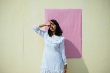 Sarı duvarın önünde duran beyaz elbiseli bir kız güçlü güneş ışığının altında pembe duvar kâğıdıyla.