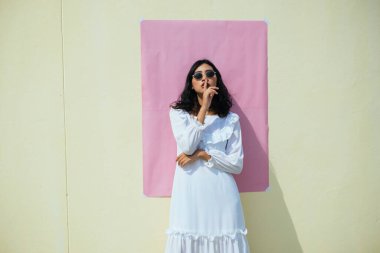 Sarı duvarda pembe kağıtlarla duran Taylandlı kız, indeksi ağzına dokunuyor..