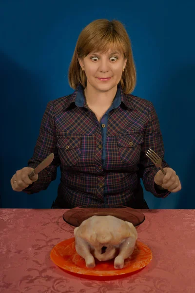 La mujer trata de comer un pollo crudo. Se sienta en una mesa con un cuchillo y un tenedor, enchufe. En un plato un pollo. Propaganda un rawismo . — Foto de Stock