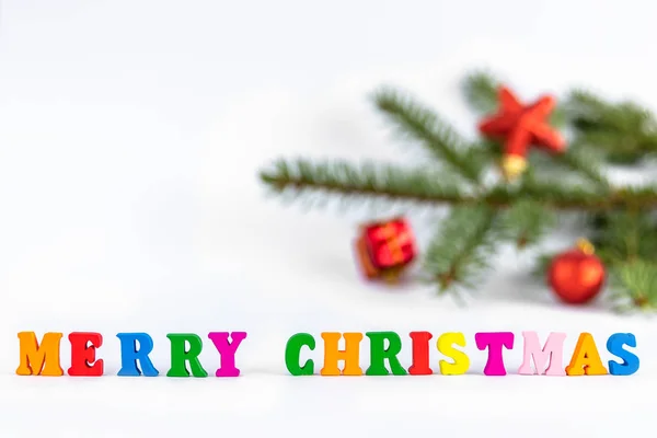 Creative layout of words merry christmas made of wooden multi-colored letters on white background with blurred fir branch and red toys. New Year concept with copy space for note and text