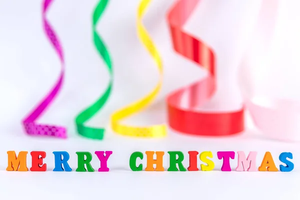 Creative layout of words merry christmas made of wooden multi-colored letters on white background with blurred curly satin ribbons. New Year concept with copy space for note and text