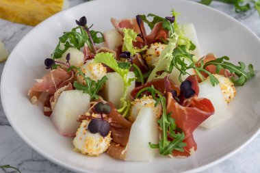 Beyaz bir tabakta kavun, proshuto, arugula ve İtalyan peyniri ile salata. Kaşık, gri mermer arka plan closeup üzerinde çatal