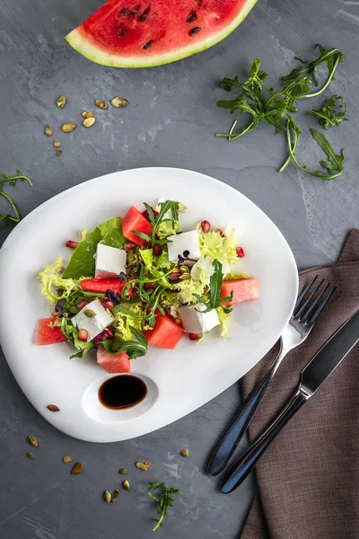 Salade d'été avec salade de pastèque et fromage feta dans une assiette blanche. Nappe marron, cuillère, fourchette sur fond gris — Photo