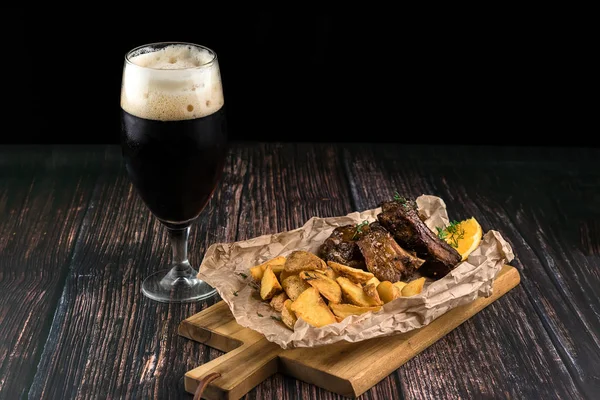 Pork ribs in honey mustard glaze with potatoes served on a wooden board and a glass of dark beer. Food concept of snack and beverages for oktoberfest beer festival with copy space