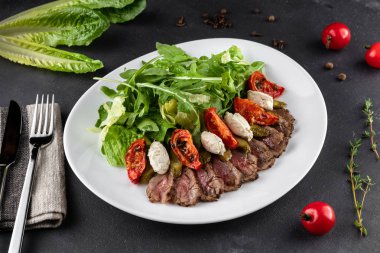 Güneşte kurutulmuş domatesli biftek salatası. Arugula, domates ve salatalık gri arka planda beyaz tabakta.