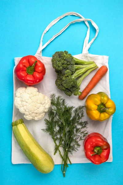 Эко-сумка из белого хлопка со свежими овощами, перцем, цветной капустой, брокколи, паччини, парсли, отходами на синем фоне с пространством для копирования — стоковое фото