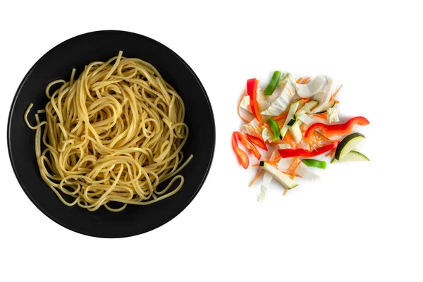 Yellow yakisoba noodles in black matte plate and colorful chopped fresh vegetables mix isolated on a white background, top view — Stock Photo, Image