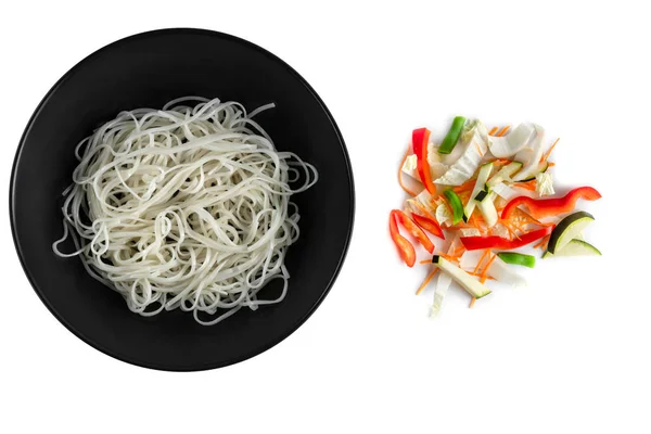 Rice noodles in black matte plate and colorful chopped fresh vegetables mix isolated on a white background, top view — Stock Photo, Image
