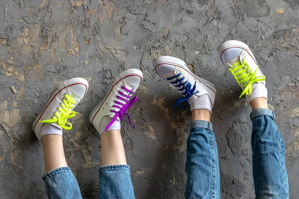 Dois pares de pernas de uma menina e um jovem vestindo jeans azul e tênis branco, pés para cima em uma parede de fundo texturizado vintage, cadarços de cor verde, rosa e azul — Fotografia de Stock