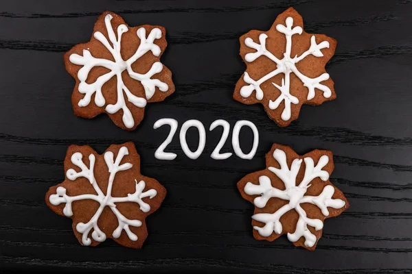 Konsep mininalistik Natal dan tahun baru pada latar belakang kayu hitam dengan angka 2020 dibuat dengan icing putih dan kue gingerbread buatan sendiri sebagai serpihan salju, pemandangan atas — Stok Foto