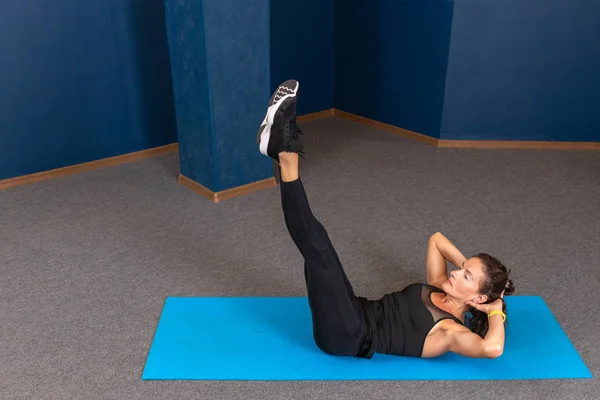 Sterke en sportieve fitnessvrouw in zwarte sportkleding die buikoefeningen doet met de handen achter het hoofd op een blauwe yogamat in de sportschool. Sport motivatie concept met Kopieerruimte voor tekst — Stockfoto