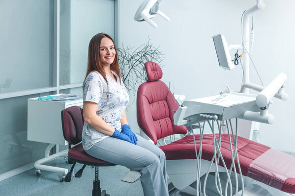 attractive good-looking portrait of female dentist in clinic sitting on chair near dental unit with hands in blue gloves, smiling and ready to accept a new patient in clinic, healthy teeth concept
