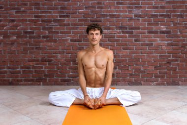 Sportif adam stüdyoda hatha yogası yapıyor. Beyaz pantolonlu bir erkek meditasyon yapıyor ve ayaklarını sarı hasırda tutuyor. Kahverengi tuğla arkaplanda kopyalama alanı olan konsept