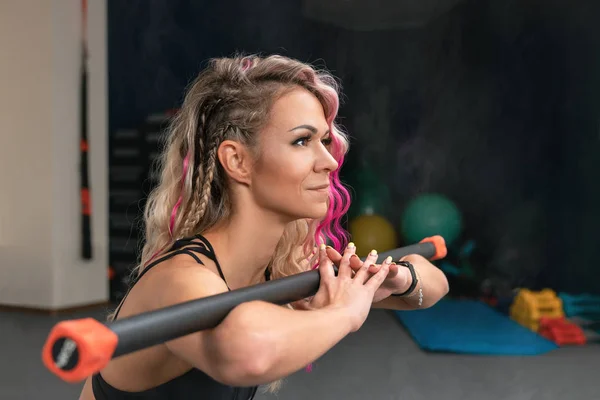 Close-up portrait of fitness woman with blond and pink long hair doing squats exercise with gymnastic stick. Blurred background of gym with copy space