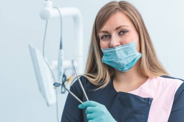 Mavi eldivenli kadın dişçi elinde bir ağız aynası ve periodontal kaşif pulu ile kliniğin arka planını diş hekimliği ve diş bakımı ile fotokopi alanı ile bulanıklaştırıyor.