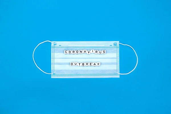 minimalist flat lay composition with white medical mask and coronavirus outbreak letters on blue color medical background, concept of protection against flu, viruses and other diseases, copy space