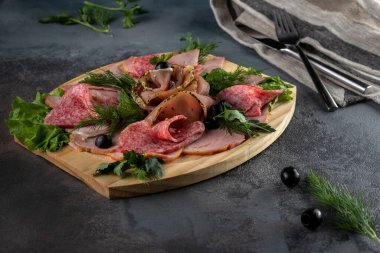 Salam, pastırma ve jambon gibi dilimlenmiş lezzetli et karışımı. Maydanozlu ve zeytinli. Masa örtüsü ve çatal bıçaklı, gri çimento arka planda tahta kesme tahtası.