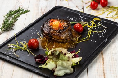 Yağlı, baharatlı ızgara biftek, siyah taş kesme tahtasında salata ve kiraz domatesleri, kırsal alanda beyaz ahşap arka planda ulusal yemek restoranının öğle yemeği menüsü.