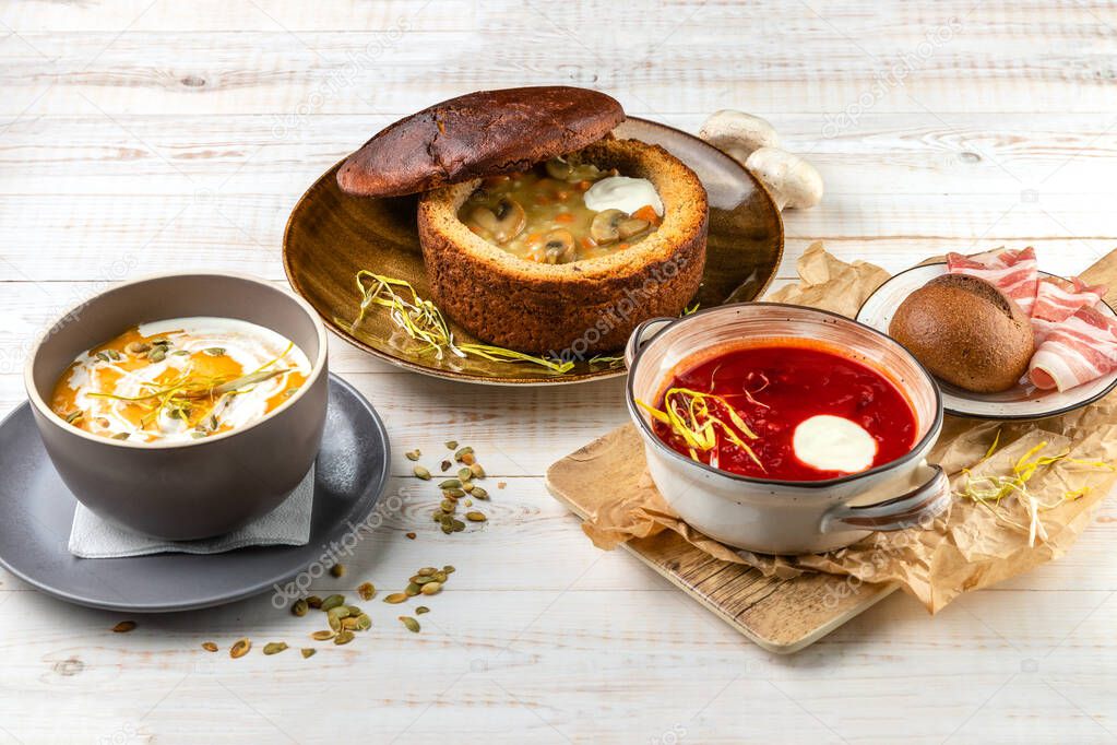 lunch menu served on white wooden background in rustic style, mushroom soup with vegetables, sour cream in bread bowl, vegetarian cream soup with pesto sauce and pumpkin seeds, red borscht with bacon