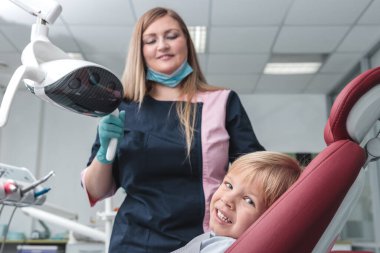 Maskeli mutlu kadın dişçi portresi, elinde dişçi aletleri, gülümseyen ve dişçi koltuğunda genç bir çocuğu muayene eden sağlıklı diş konsepti.