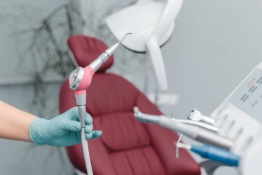 Mavi eldivenli dişçinin kadın eli, elinde kum püskürtme aleti, kliniğin arka planında bulanıklık, diş hekimliği ve fotokopi alanı olan diş bakımı kavramı.
