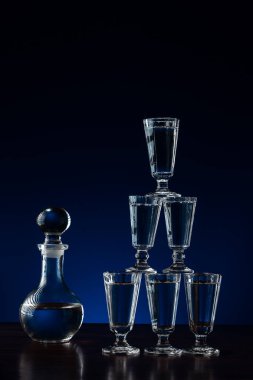 Kristal sürahi veya bir sürahi votka ve altı bardak piramit kulesi olarak restorandaki ahşap masa üzerine yerleştirilmiş, mavi ve siyah arkaplan ve fotokopi alanı