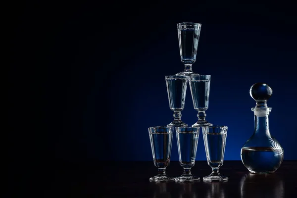 Crystal decanter or carafe of vodka and glasses pyramid tower on wooden table in restaurant, blue and black background with copy space