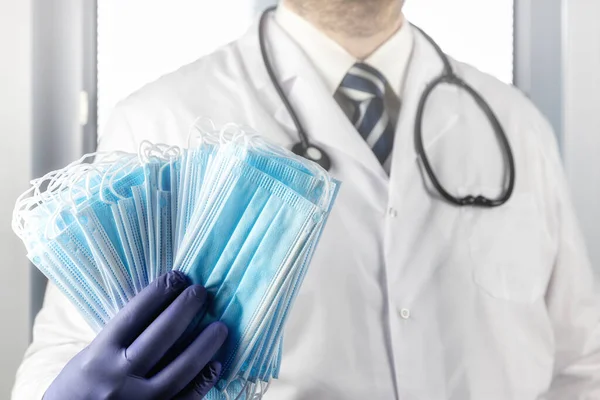 Lekarz Białym Fartuchu Stetoskopem Rękawiczkami Trzyma Stos Niebieskich Masek Medycznych — Zdjęcie stockowe