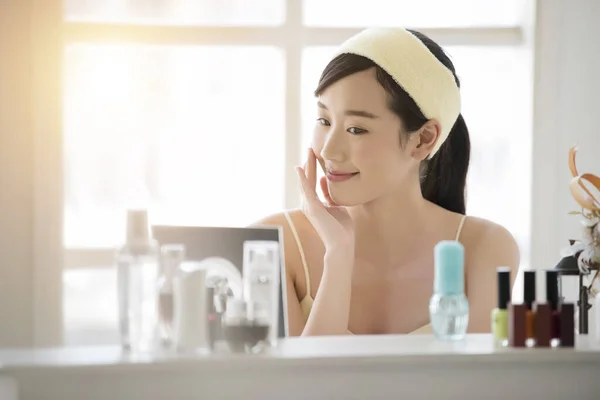 Smiling young beautiful Asian woman fresh healthy skin looking on mirror with serum and lotion set at home.facial beauty and cosmetic concept