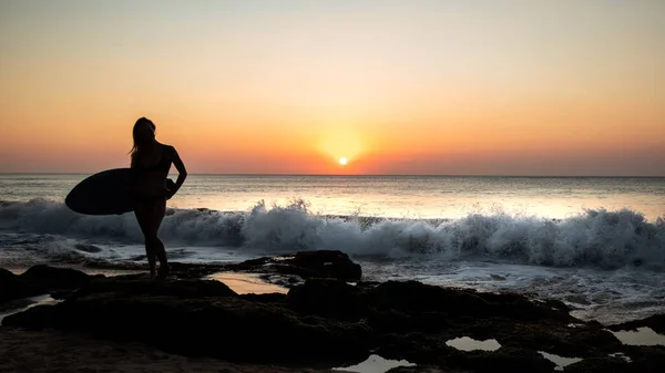 Plajda sörf tahtası olan sörfçü kız silueti. Gün batımı zamanı. Tegal Wangi Plajı, Bali, Endonezya