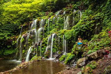 Genç bir kadın kayanın üzerinde oturuyor, şelalenin yanında yoga yapıyor. Eller Gyan Mudra 'da. Banyu Wana Amertha şelalesi, Bali. Arkadan görüntüle.