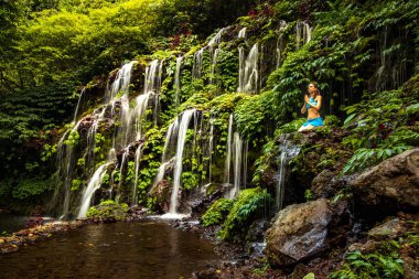 Kayanın üzerinde oturan bir kadın, şelalenin yanında yoga ve pranayama yapıyor. Eller namaste mudra 'da. Banyu Wana Amertha Şelalesi, Bali. 
