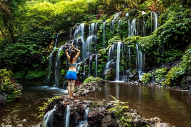 Kayanın üzerinde durmuş yoga yapan bir kadın. Şelale yakınlarında namaste Mudra ile kol kaldıran genç bir kadın. Banyu Wana Amertha şelalesi, Bali. Arkadan görüntüle.