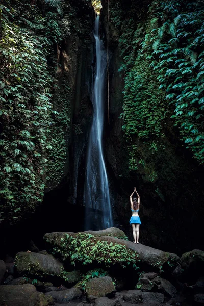 Женщина, стоящая на камне, практикующая йогу. Молодая женщина поднимает руки с мудрой намасте возле водопада. Водопад Леке Леке, Бали. Вид со спины . — стоковое фото