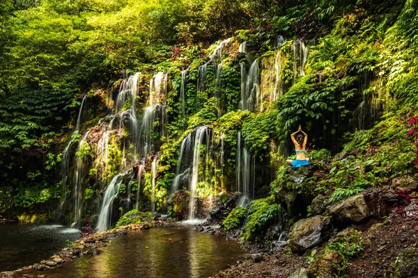 Woman sitting on the rock, practicing yoga. Young woman raising arms with namaste mudra near waterfall. Banyu Wana Amertha waterfall, Bali. View from back. — Stock Photo, Image