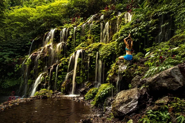 Femme assise sur le rocher, pratiquant le yoga. Jeune femme levant les bras avec namaste mudra près de cascade. Cascade de Banyu Wana Amertha, Bali. Vue de dos . — Photo