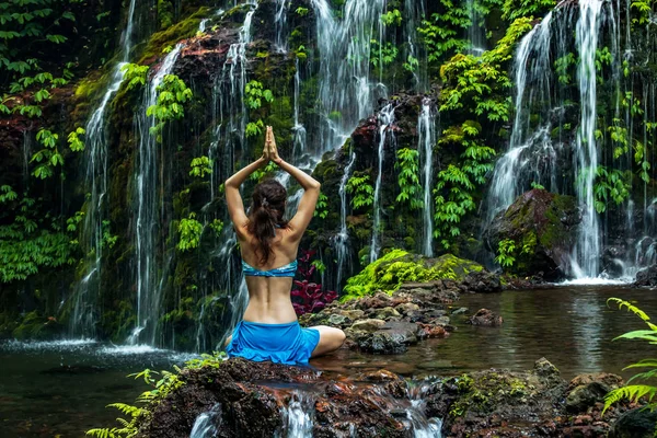 Kvinnan sitter på klippan och utövar yoga. Ung kvinna höjer armarna med namaste mudra nära vattenfallet. Banyu Wana Amertha vattenfall, Bali. Utsikt bakifrån. — Stockfoto