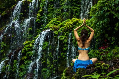 Kayanın üzerinde oturan bir kadın, yoga yapıyor. Şelale yakınlarında namaste Mudra ile kol kaldıran genç bir kadın. Banyu Wana Amertha şelalesi, Bali. Arkadan görüntüle.